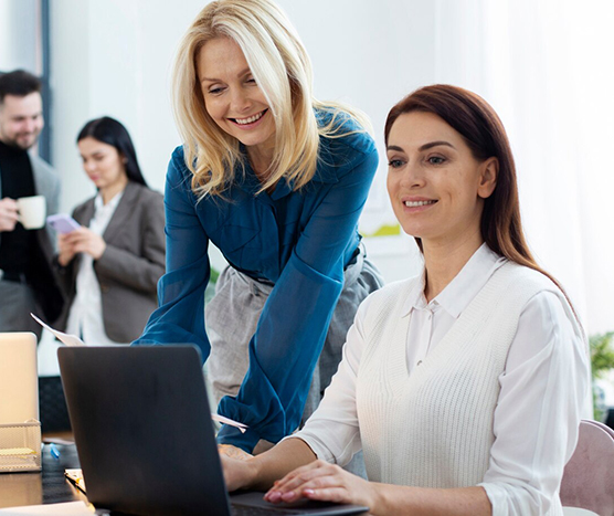 Two women working at a desk, one standing and smiling, the other seated and typing on a laptop.